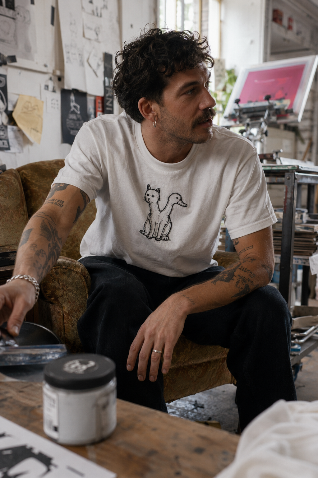 Man wearing a white Trees, Rivers & UFOs Quackcat T-shirt while sitting forward in a cluttered creative studio.