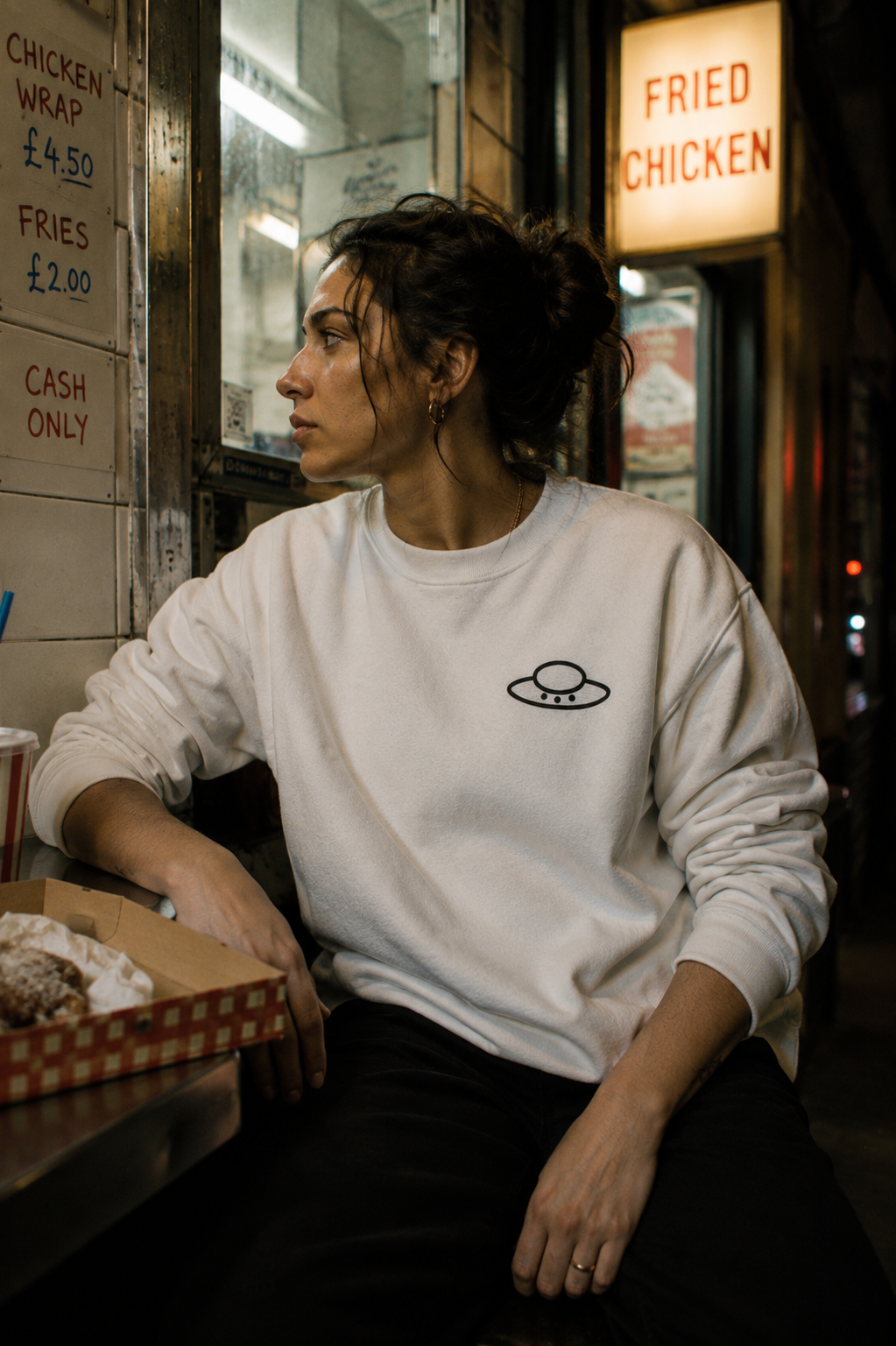 Woman wearing a white embroidered UFO sweatshirt while sitting inside a late-night fried chicken shop.