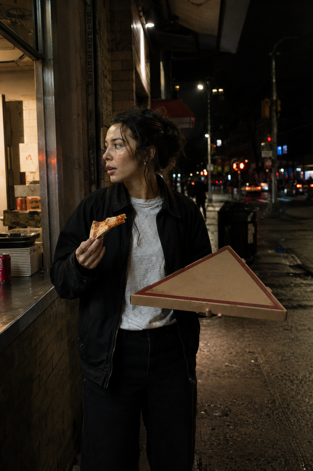 Woman eating pizza outside a late-night takeaway while holding an ordinary-looking triangular pizza box horizontally.