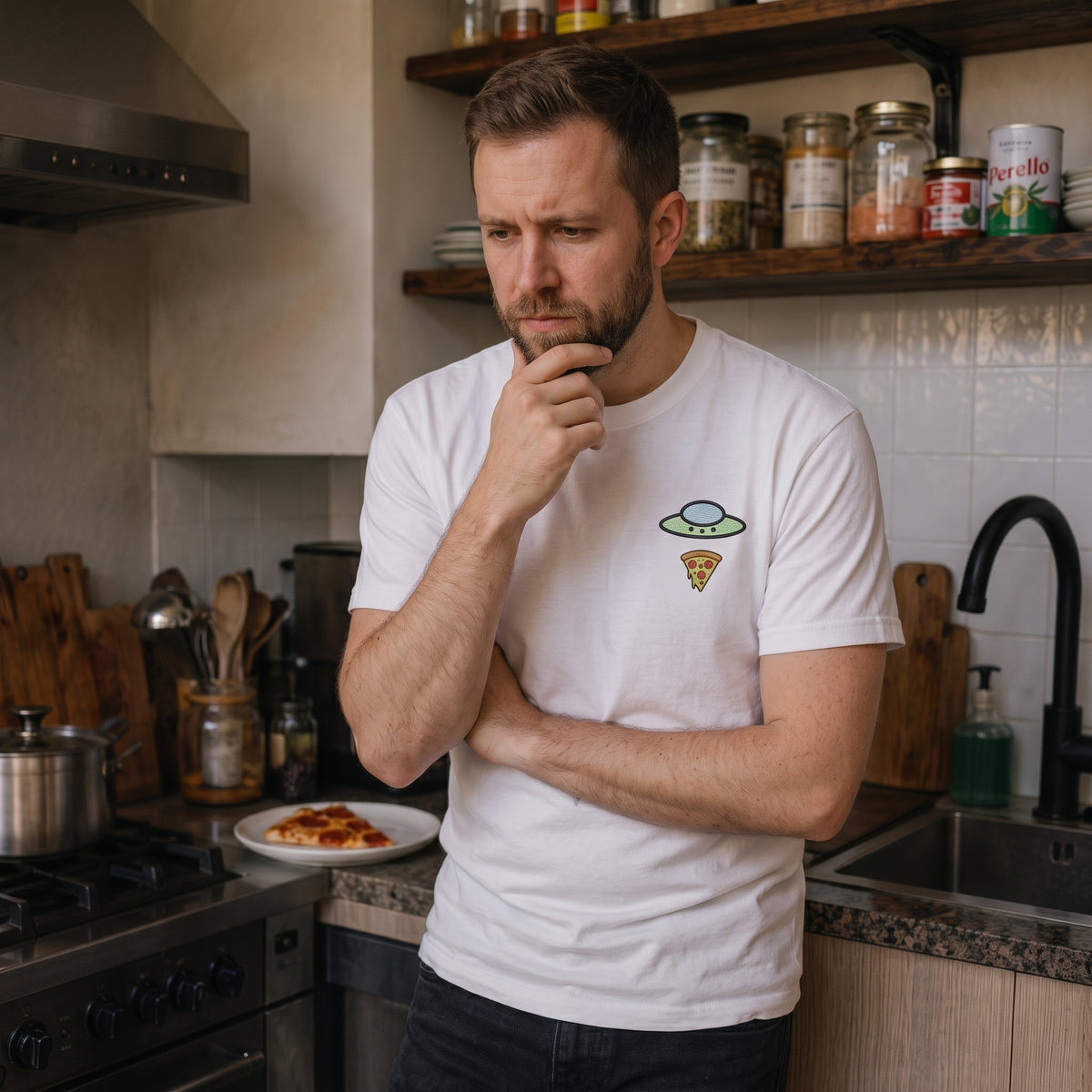 Man wearing the white Trees, Rivers & UFOs Forest Pizza Tee in a kitchen with his hand on his chin.