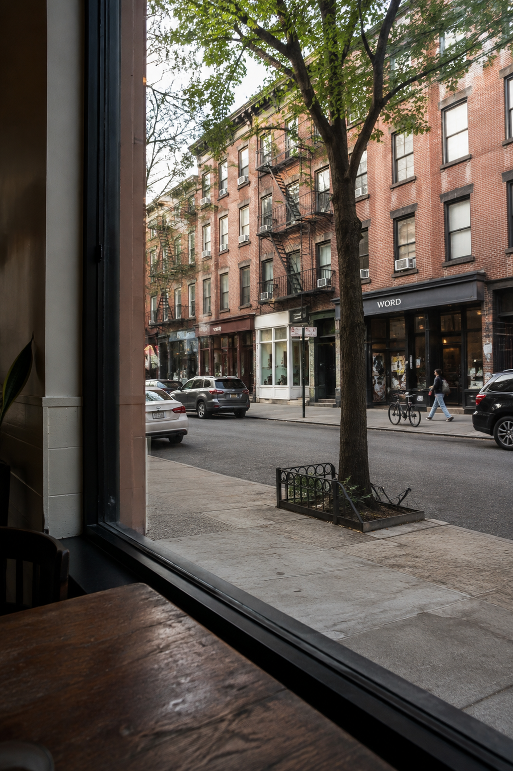 A view from inside a cafe or restaurant looking out onto a city street with brick apartment buildings, parked cars, a tree, and a sidewalk.