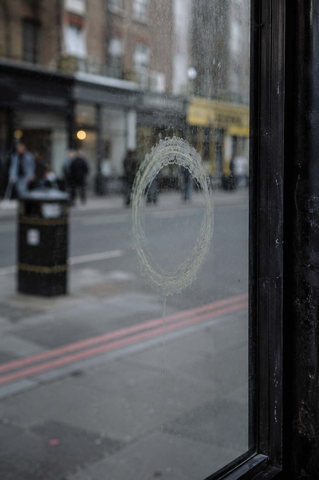 A close-up of a dirty, smoky glass window pane with a circular scratch or impact mark in the center, looking out onto a blurred city street with pedestrians and buildings.