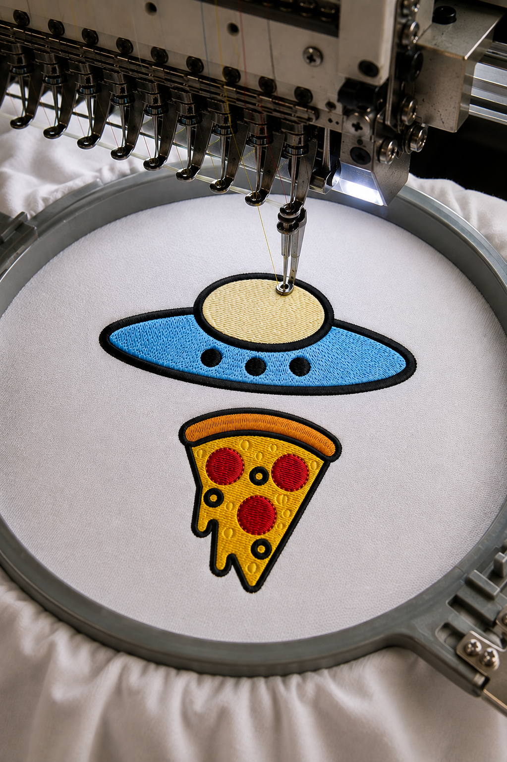 Embroidery machine stitching a blue UFO above a yellow pizza slice on white fabric.
