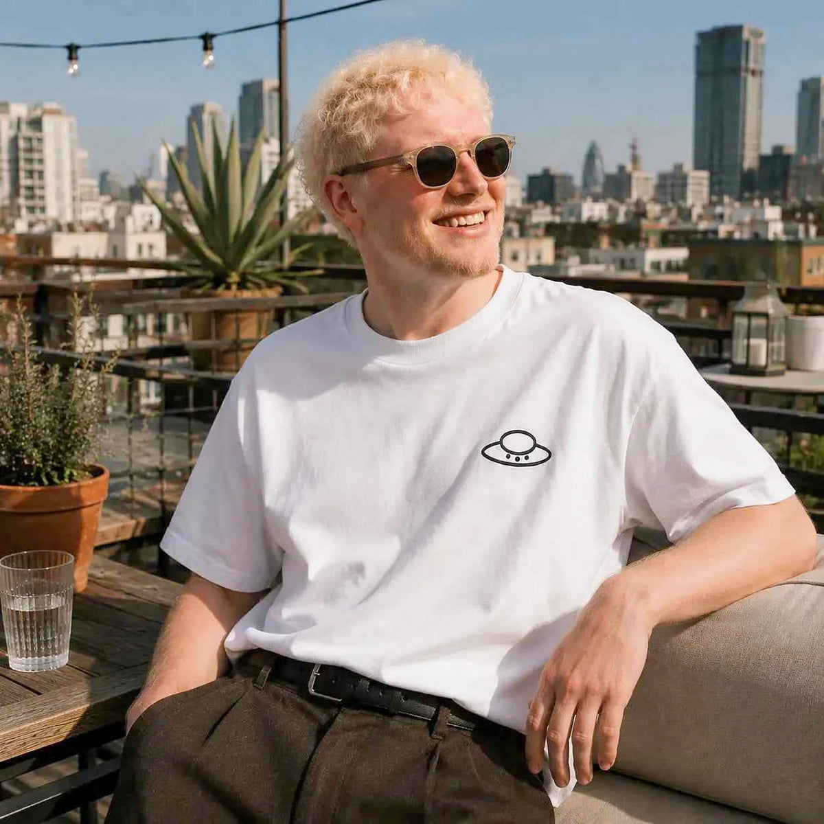 Man wearing the white Trees, Rivers & UFOs Nimbus Tee with brown trousers and sunglasses while seated on a rooftop terrace.