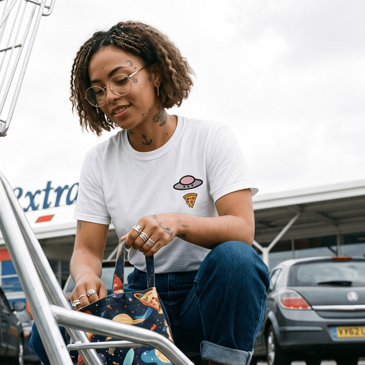 Woman wearing the white Trees, Rivers & UFOs Orchid Pizza Tee with blue jeans while seated in a parking lot and holding a patterned tote bag.