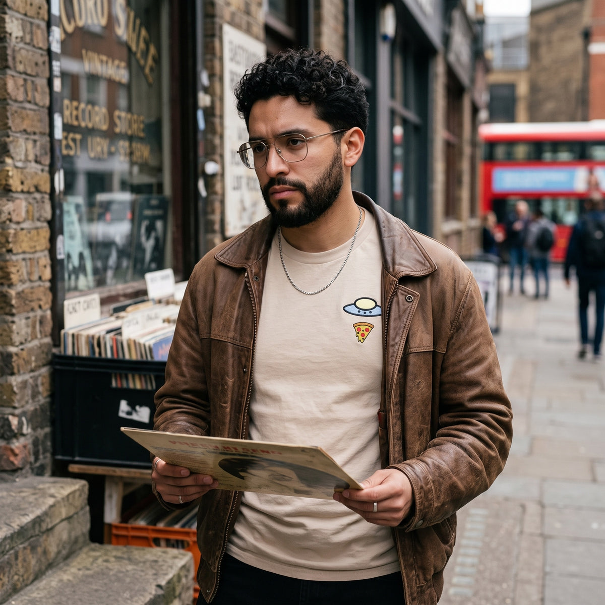 A man wearing a brown leather jacket over a beige t-shirt with a small pizza and alien graphic, standing on a city street and holding a vinyl record.