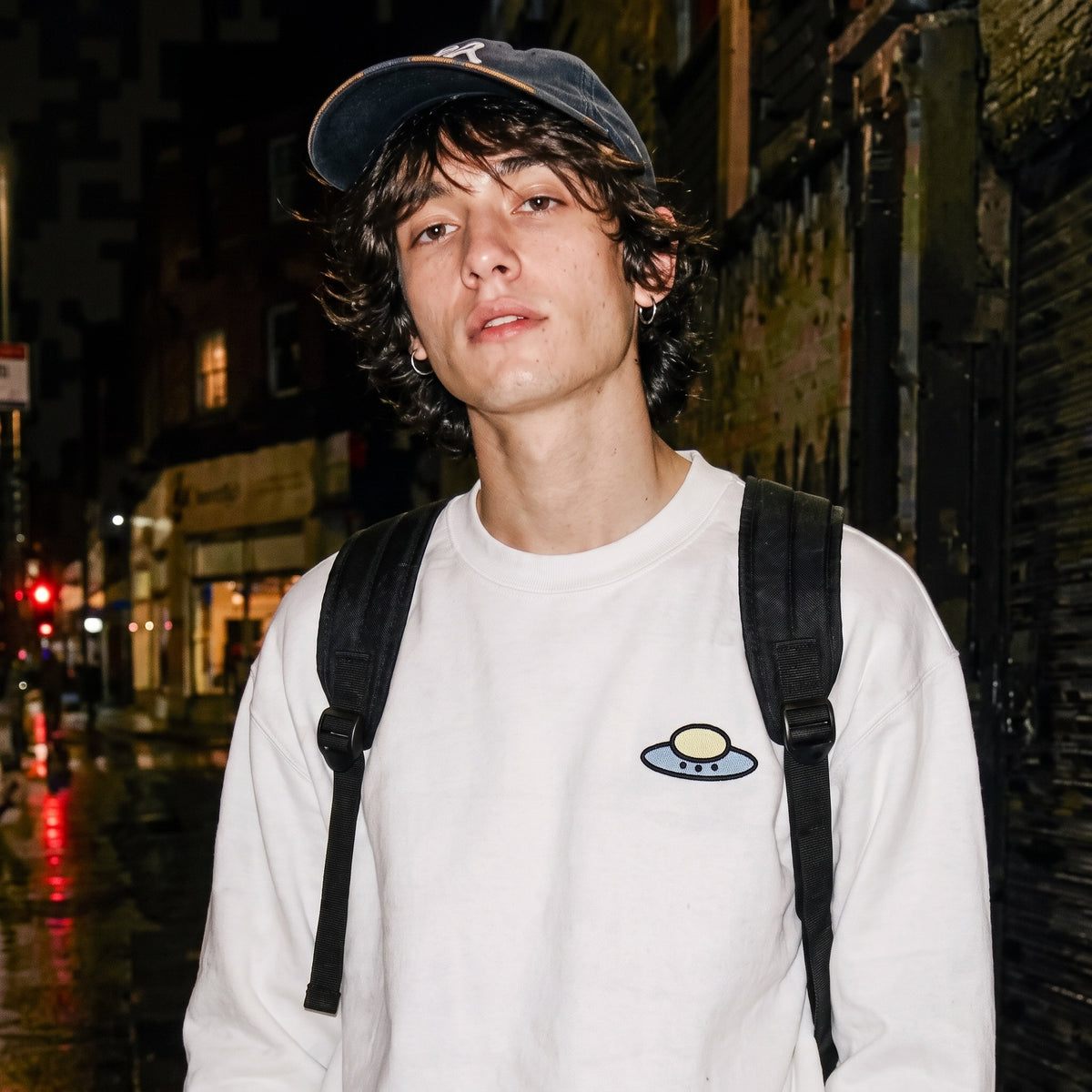 Man wearing the white Trees, Rivers & UFOs Sandstone Sweatshirt with a dark cap and backpack on a wet city street at night.