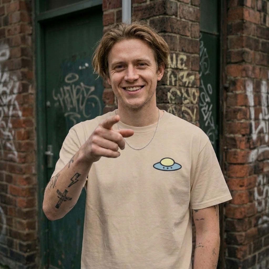 A smiling man with tattoos points directly at the camera while wearing a beige crewneck t-shirt with a small blue and yellow UFO graphic on the left chest, standing in front of a brick wall and a green graffiti-covered door.