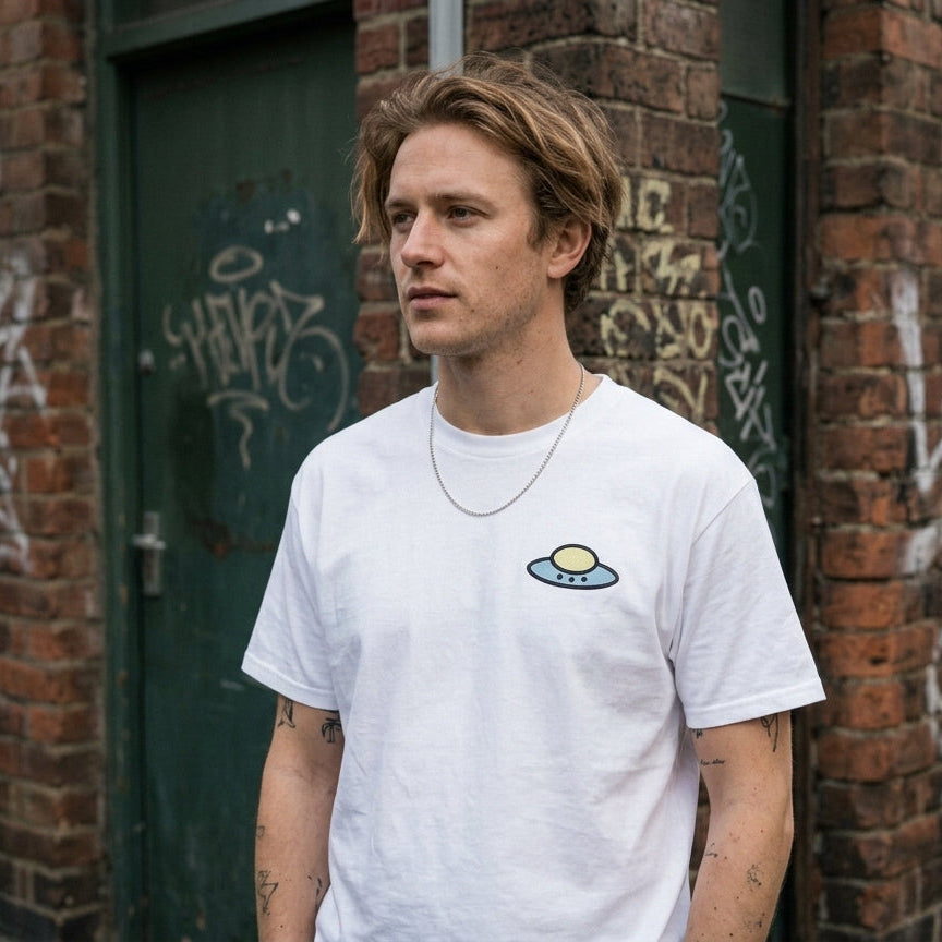 A man wearing a white crewneck t-shirt with a small yellow and blue alien graphic on the left chest, standing in front of a brick wall with graffiti.