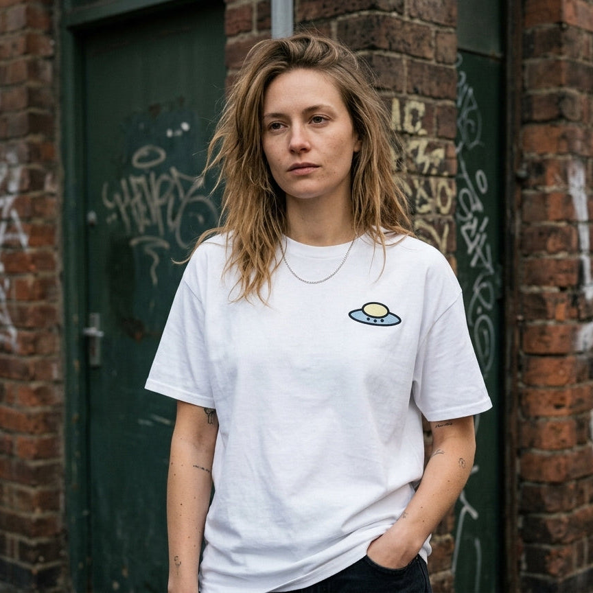 A woman with light brown hair stands in front of a graffiti-covered brick wall wearing a white crewneck t-shirt with a small cartoon planet graphic on the left chest.
