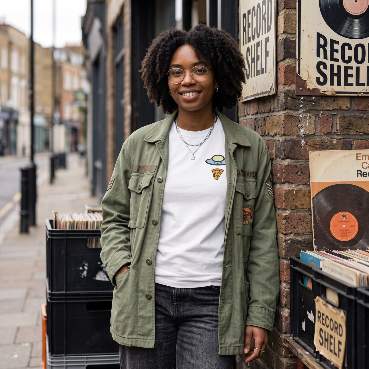 A woman with curly hair and glasses stands on a city street wearing an olive green utility jacket over a white graphic t-shirt with a UFO design, paired with dark gray jeans. She is positioned next to a brick wall decorated with vintage 'Record Shelf' signs and crates of vinyl records.